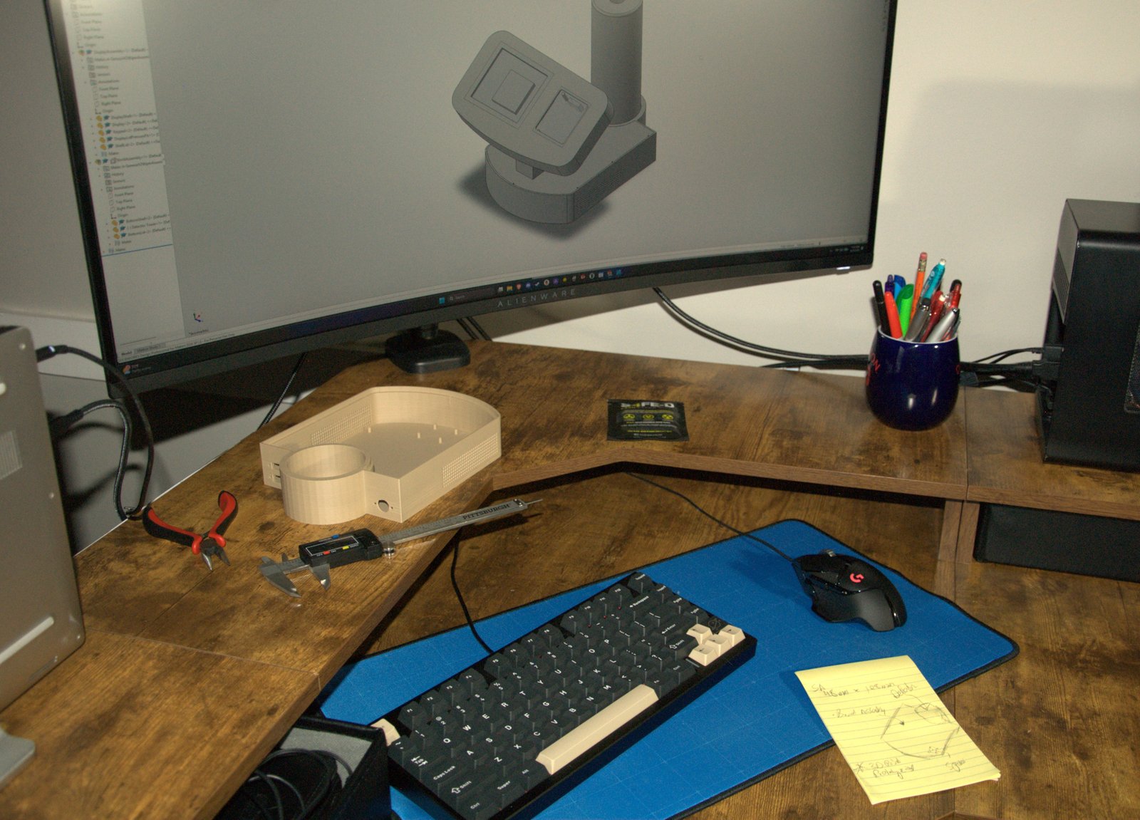LTI engineering workstation — CAD design of a new gamma-counter housing on the ultrawide monitor, a 3D-printed prototype on the bench, calipers and pliers within reach, and an S4FE-D D.I.R.T. wipe pouch on the shelf — both product lines on one bench