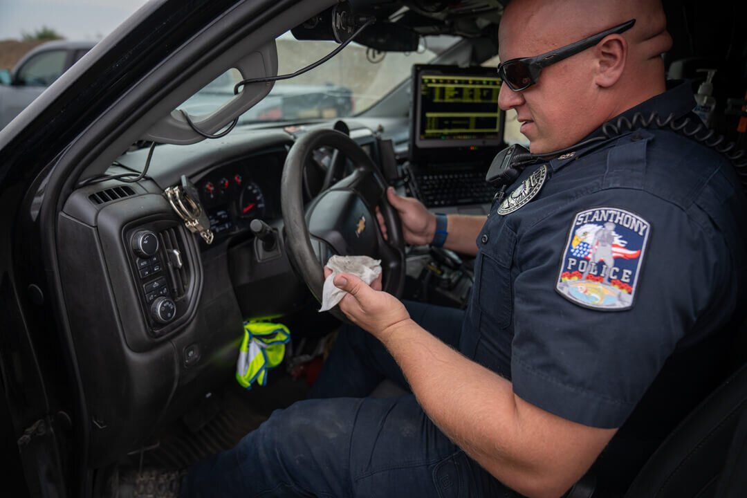 St. Anthony Police officer sitting in the driver's seat of a patrol unit, using an S4FE-D® D.I.R.T. wipe to clean the steering wheel and dash &mdash; routine cruiser-interior decon after a callout.
