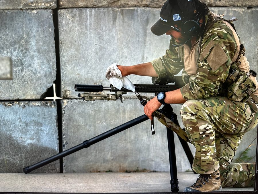 Operator wiping down a precision rifle upper receiver with an S4FE-D® D.I.R.T. wipe at the range — multicam BDUs, ear protection, real post-shift weapon cleanup.