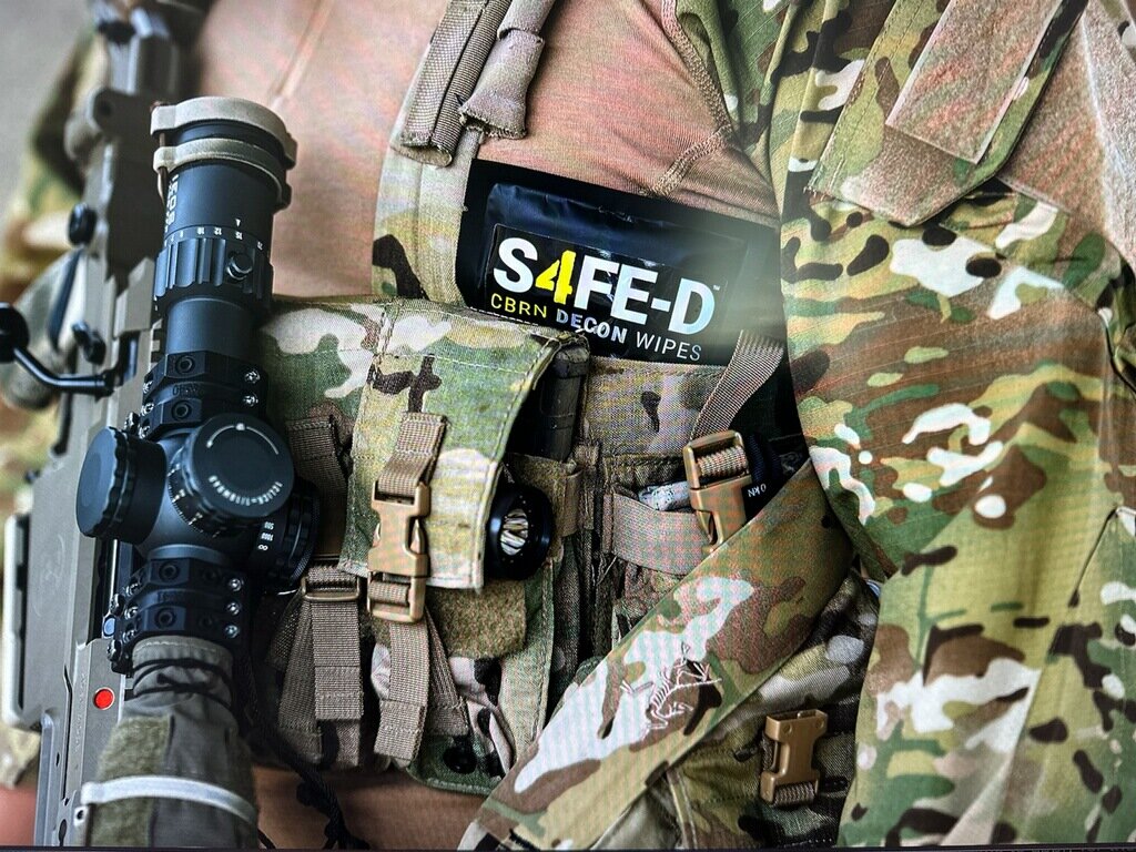 Close-up of a tactical operator's MultiCam plate carrier with an S4FE-D® CBRN Decon Wipes pouch stowed in a front placard pouch on the carrier, alongside a precision rifle — wipe is pre-positioned on the body, ready to grab.