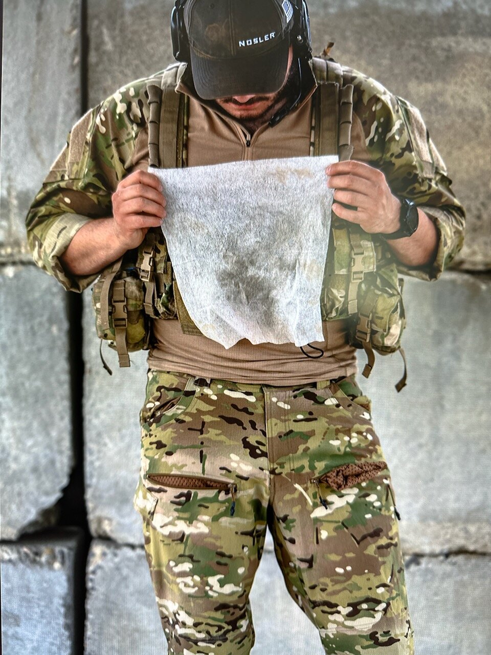 Same operator after the rifle and optic wipe-down, holding up the used S4FE-D® D.I.R.T. wipe in front of him — the wipe is visibly streaked and stained with the lead, primer residue, carbon, and lubricant the chemistry lifted off the rifle and suppressor.