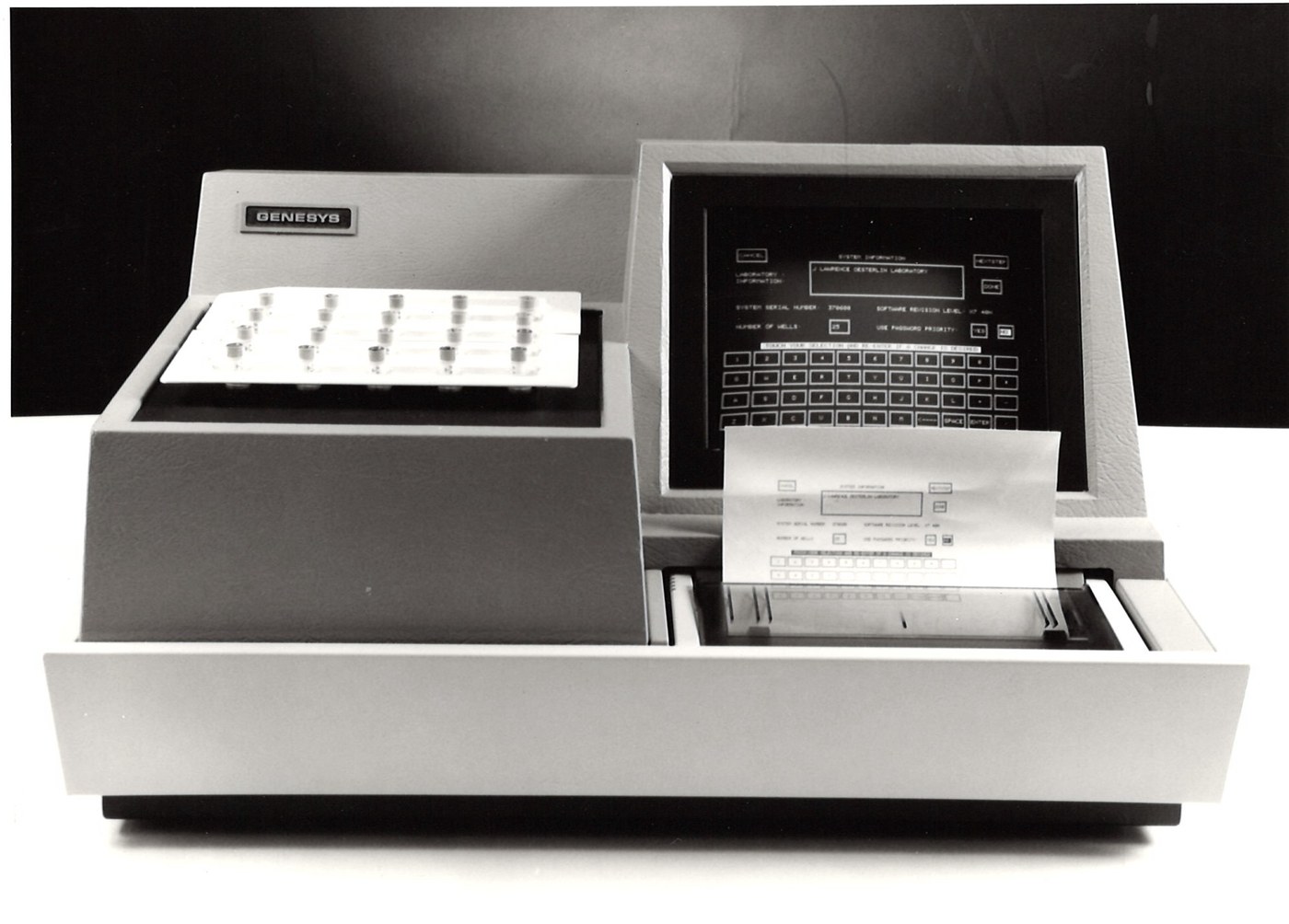A Genesys 5000 multi-well gamma counter — the LTI flagship of the late 1980s and 1990s. Angled CRT display on the right with on-screen virtual keypad, external sample tray on the left holding a grid of sample tubes, paper output emerging from the front printer port, GENESYS badge visible on the left side.
