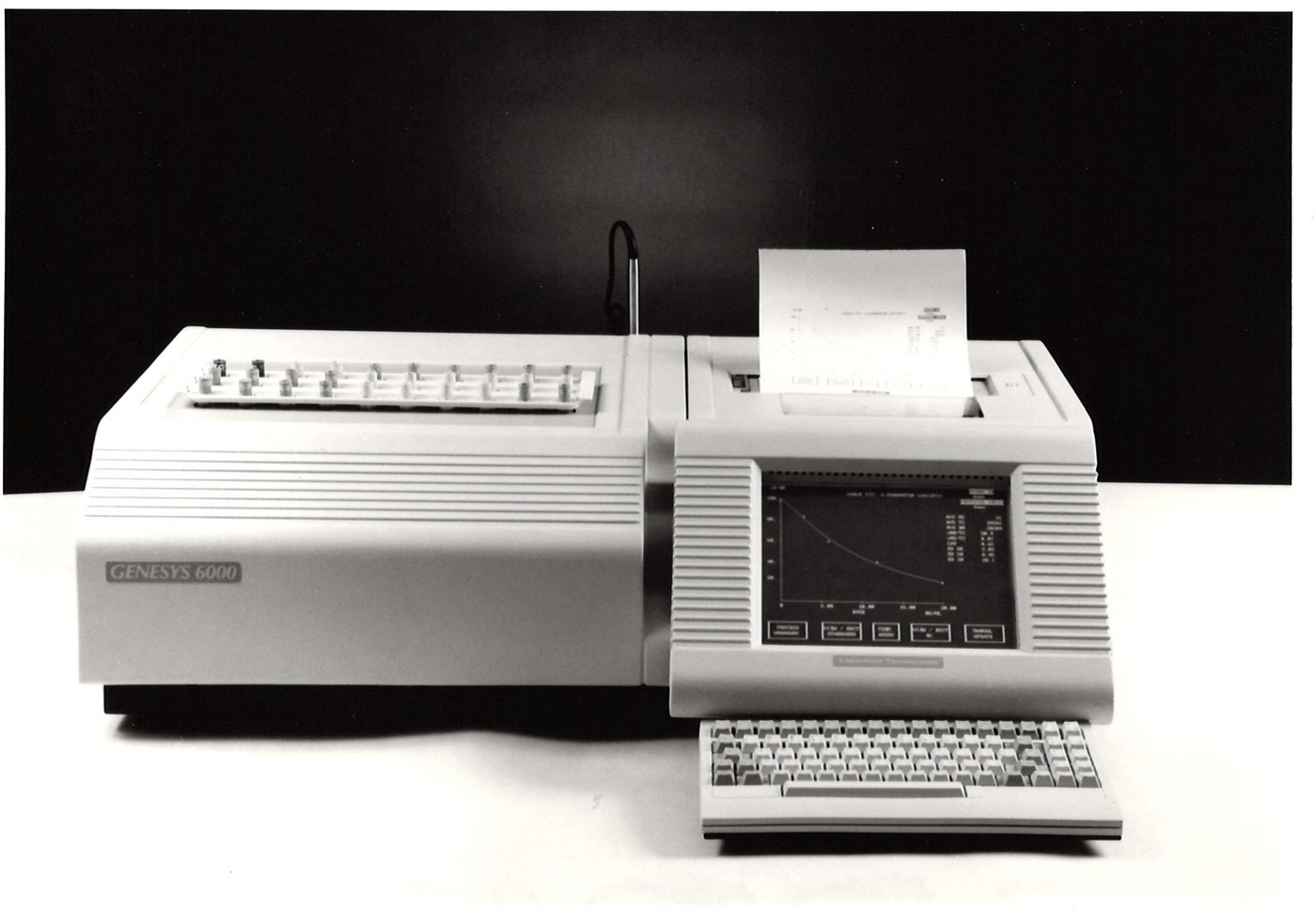 A Genesys 6000 multi-well gamma counter — visibly larger than the 5000 chassis, with a separate angled control console showing a graphical efficiency curve on the CRT and an external keyboard at the bench, plus a separate sample-loader unit on the left with a grid of sample positions. Late 1980s / early 1990s product photo.