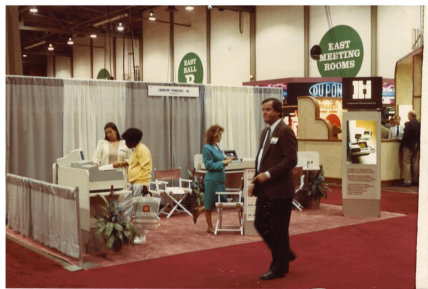 Laboratory Technologies, Inc. trade show booth at a clinical industry conference, late 1980s. Director's-chair seating, curtained backdrop, LTI signage. DuPont and other major-vendor booths visible in the background. A man in a suit walks past in the foreground.