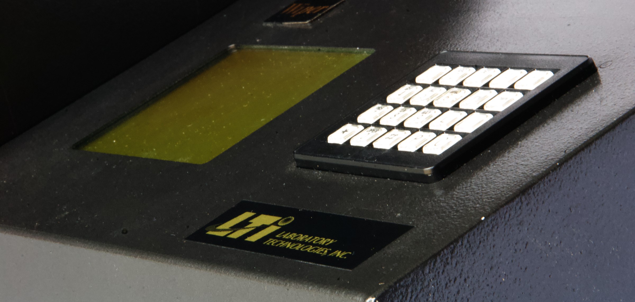 Detail of the Multi-Wiper control panel — backlit LCD with green phosphor, metallic keypad, gold LTI Laboratory Technologies, Inc. nameplate, all under low-key lighting